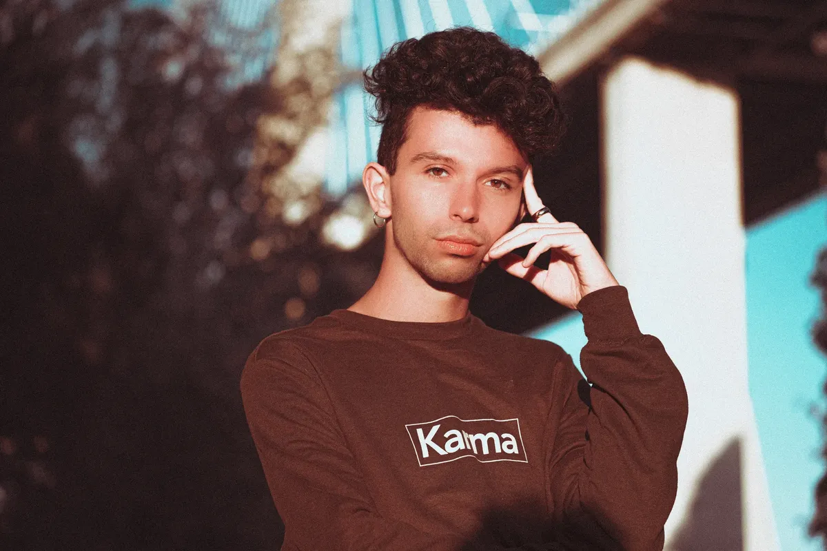 Stylish young man with curly hair posing on a sunny day, wearing a 'Karma' sweatshirt.