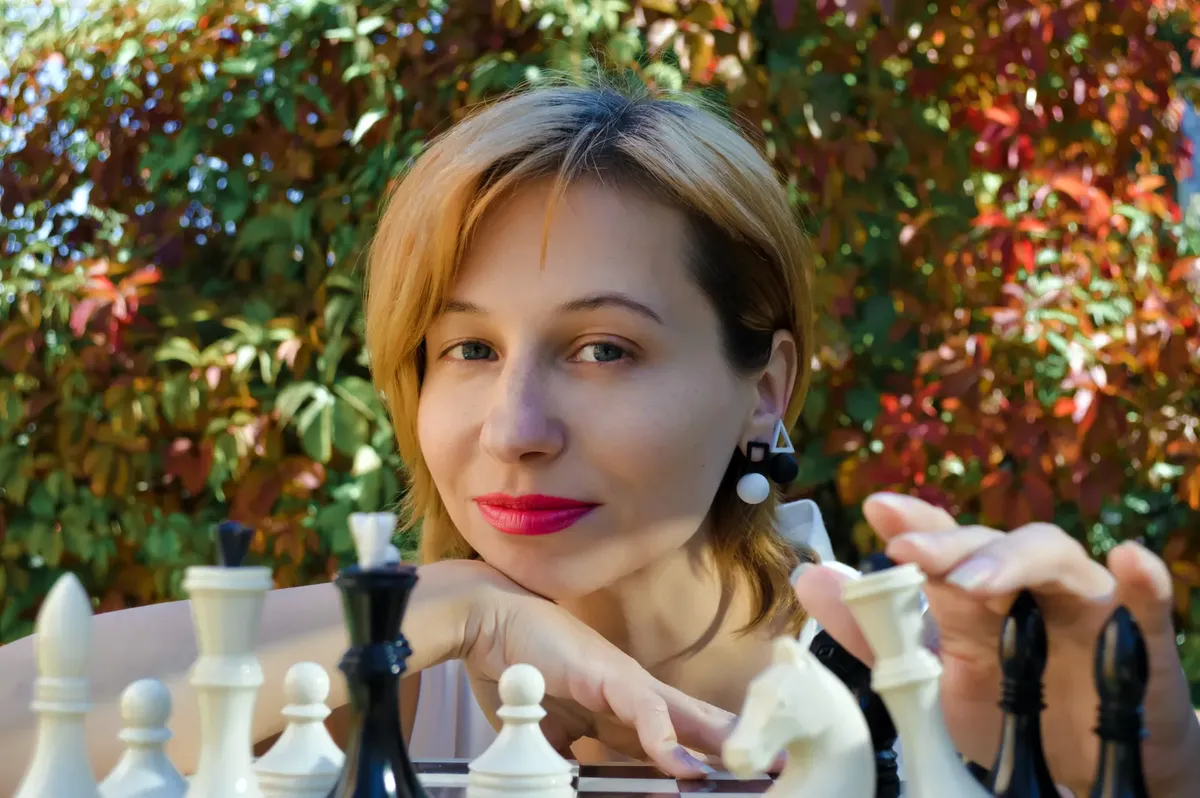 內容為女性玩家面對攝像機，坐在棋盤上，於街頭進行棋局，周圍有色彩豐富的樹木 foliage