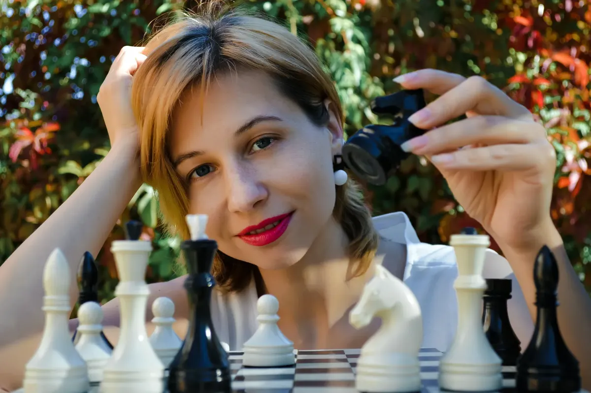 開心的年輕女性國際象棋選手，小棋子靠在手上，坐在公園棋盤旁 looking at camera