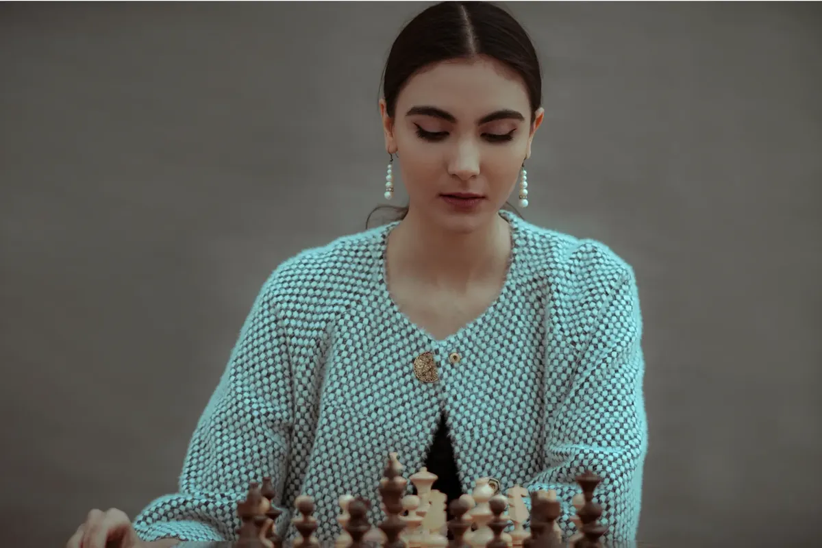Concentrated ethnic female player sitting at table and playing chess board game of strategic skill against gray background