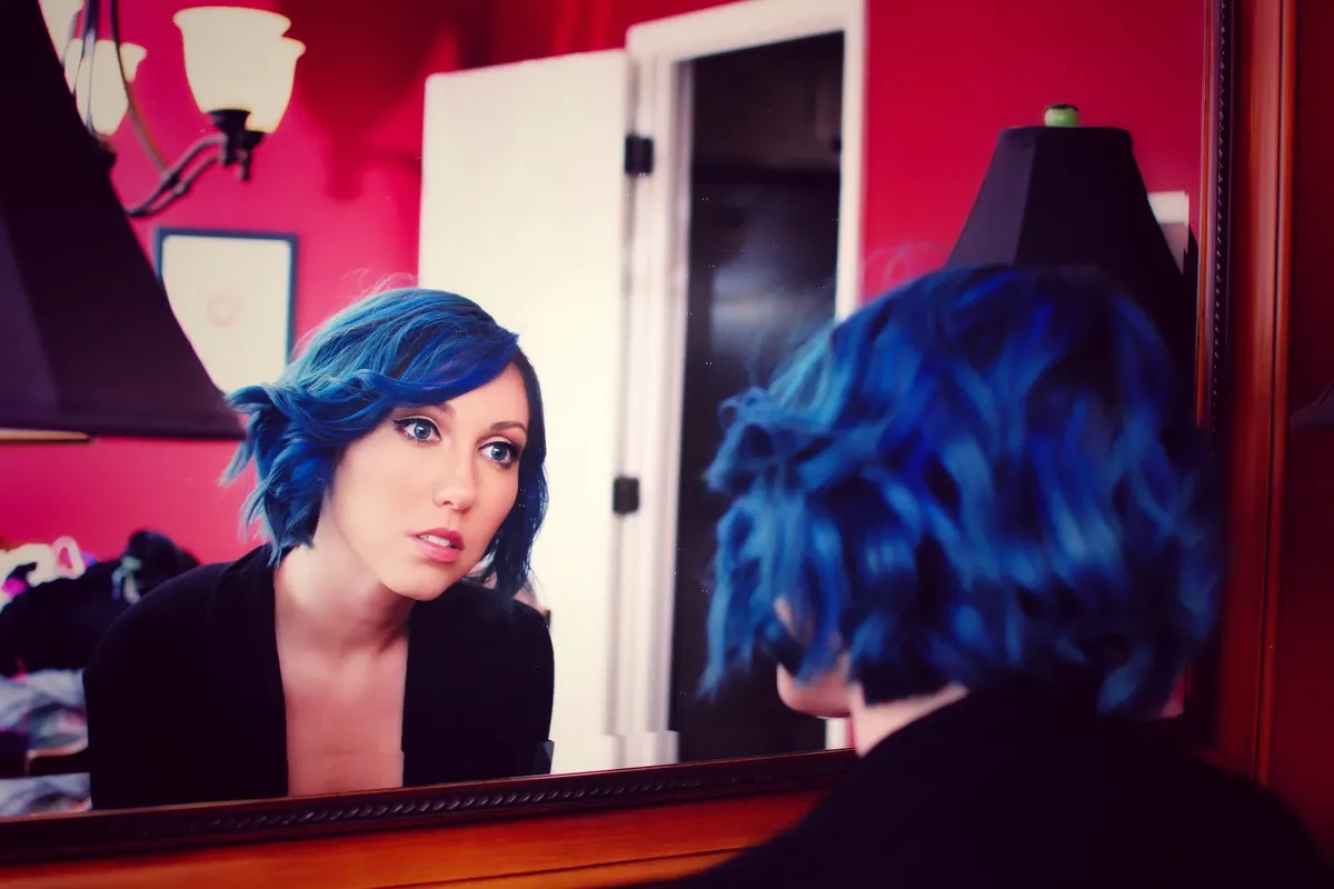 A woman with blue hair looks at her reflection in a mirror, indoors with red walls.